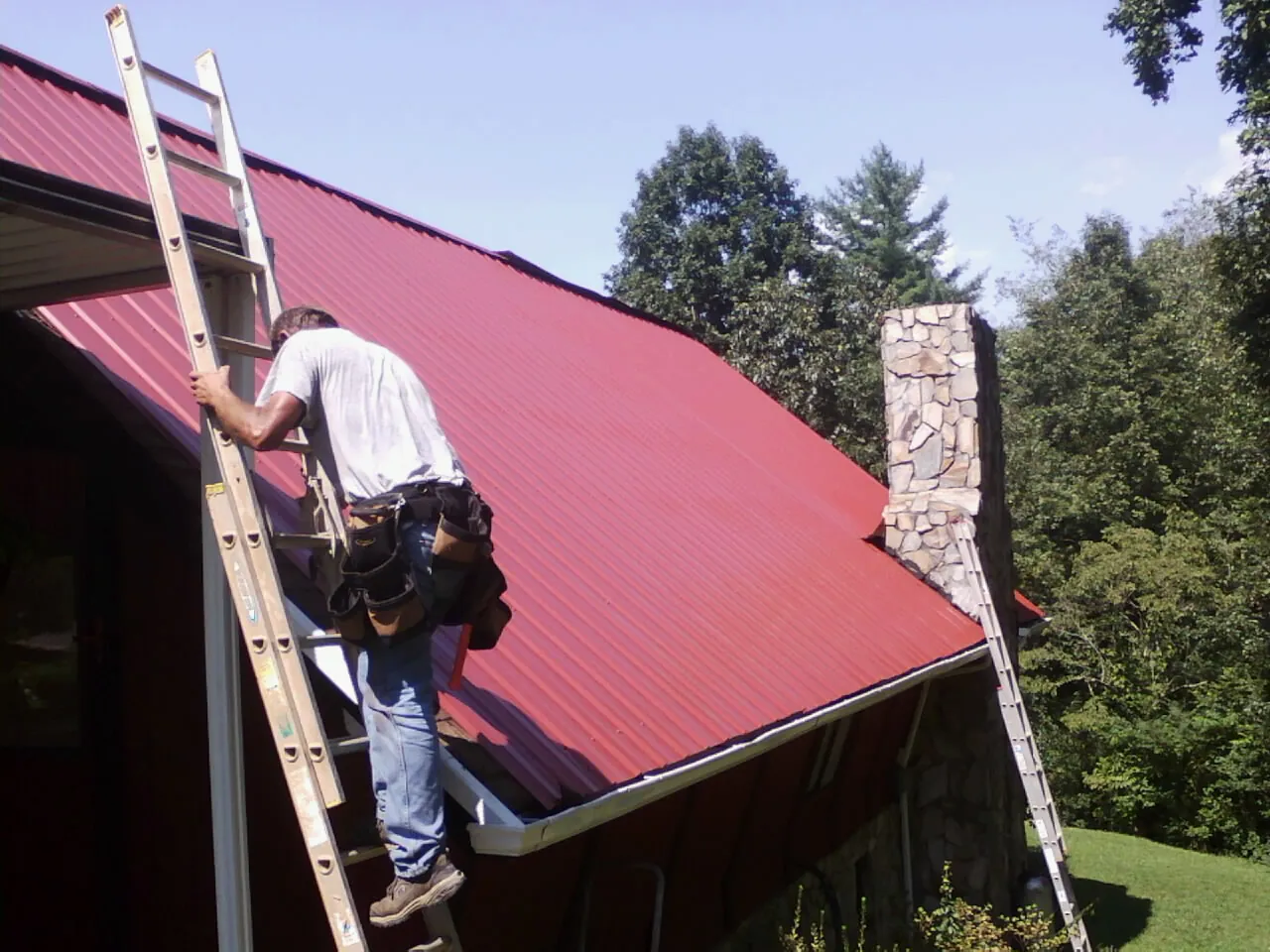 Roofer on a ladder beside a red metal roof and stone chimney — Soffit & Fascia Repair by Roofing Brothers in Mc David